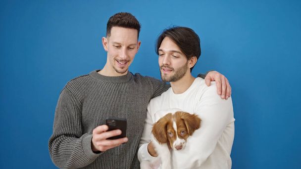 Dos hombres pareja usando teléfono inteligente celebración perro sobre aislado fondo azul - Foto, Imagen