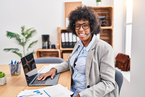 Черная женщина с вьющимися волосами в наушниках агента колл-центра в офисе подмигивает, глядя на камеру с сексуальным выражением лица, веселым и счастливым лицом.  - Фото, изображение
