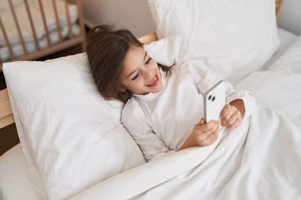 Adorable hispanic girl using smartphone lying on bed at bedroom - Fotoğraf, Görsel