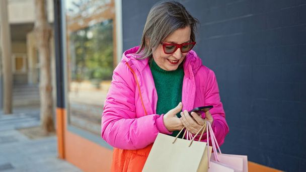 volwassen latino vrouw met grijs haar glimlachen gaan winkelen met behulp van smartphone op straat - Foto, afbeelding