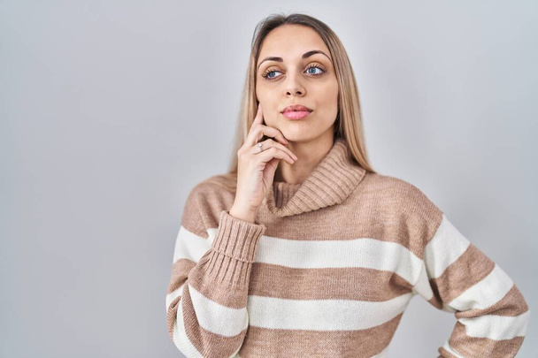若いブロンドの女性は、質問、集中的な表現について考える顎の手で孤立した背景にタートルネックセーターを着ています。思慮深い顔で笑顔。疑わしい概念.  - 写真・画像