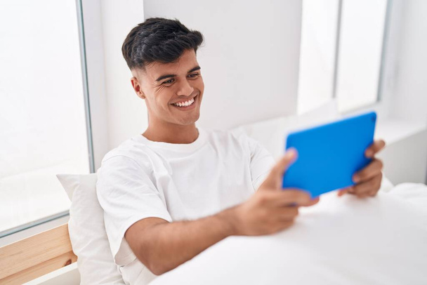 Young hispanic man using touchpad sitting on bed at bedroom - Photo, image