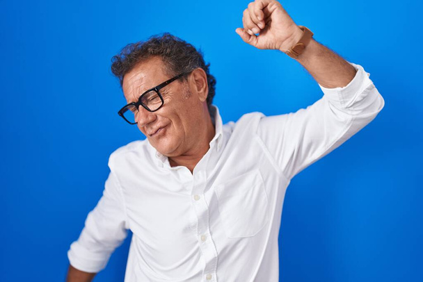 Middle age hispanic man standing over blue background dancing happy and cheerful, smiling moving casual and confident listening to music  - Fotografie, Obrázek