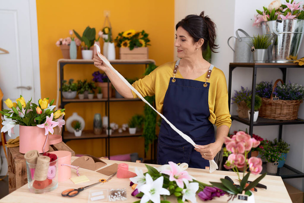Florista hispana de mediana edad sonriendo confiada sosteniendo encaje de regalo en la tienda de flores - Foto, Imagen