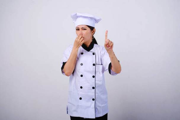 Photo of disappointed and unhappy female Asian chef standing with hand covering nose, bad smell gesture, feeling disgusted on empty space or copy space for product isolated on white background - Valokuva, kuva