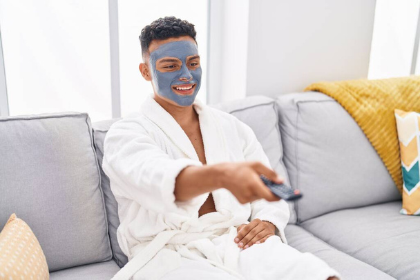 Joven latino relajado en sofá con tratamiento de mascarilla facial viendo tv en casa - Foto, Imagen