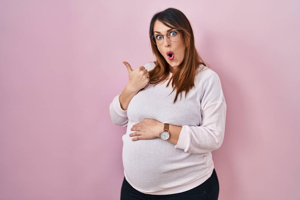 Těhotná žena stojící nad růžovým pozadím překvapeně ukazuje ukazováčkem na stranu, otevřená ústa užaslý výraz.  - Fotografie, Obrázek