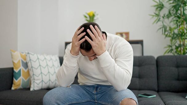Young hispanic man stressed sitting on sofa at home - 写真・画像