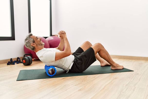 Middle age grey-haired man using foam roller stretching at sport center - Фото, изображение