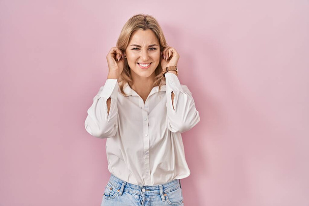 Jeune femme caucasienne portant une chemise blanche décontractée sur fond rose couvrant les oreilles avec des doigts avec une expression agacée pour le bruit de la musique forte. concept sourd.  - Photo, image