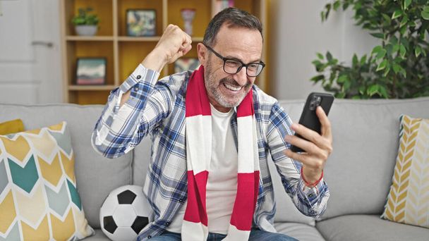 Hombre de mediana edad que apoya al equipo de fútbol viendo el juego en el teléfono inteligente en casa - Foto, Imagen