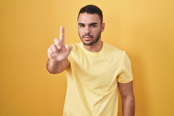 Joven hispano de pie sobre fondo amarillo señalando con el dedo hacia arriba y la expresión enojada, sin mostrar ningún gesto  - Foto, imagen