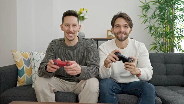 Dos hombres en pareja sentados en el sofá jugando videojuegos en casa - Foto, Imagen