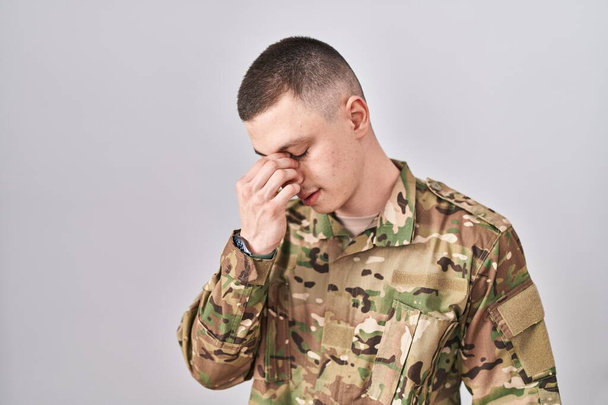 Young man wearing camouflage army uniform tired rubbing nose and eyes feeling fatigue and headache. stress and frustration concept.  - Photo, image