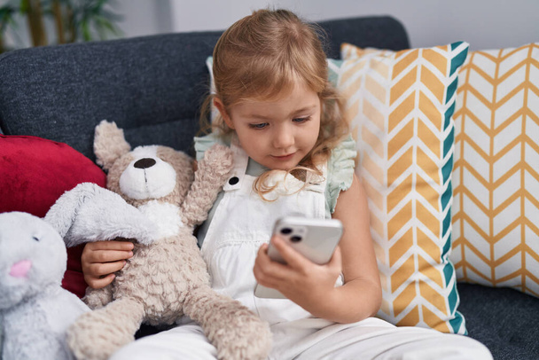 Liebenswertes blondes Mädchen mit Smartphone umarmt Teddybär zu Hause - Foto, Bild