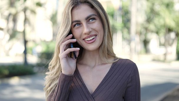 Молодая блондинка улыбается уверенно разговаривая по смартфону в парке - Фото, изображение