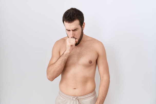 Junger hispanischer Mann, der ohne Hemd vor weißem Hintergrund steht, fühlt sich unwohl und hustet als Symptom für Erkältung oder Bronchitis. Gesundheitskonzept.  - Foto, Bild
