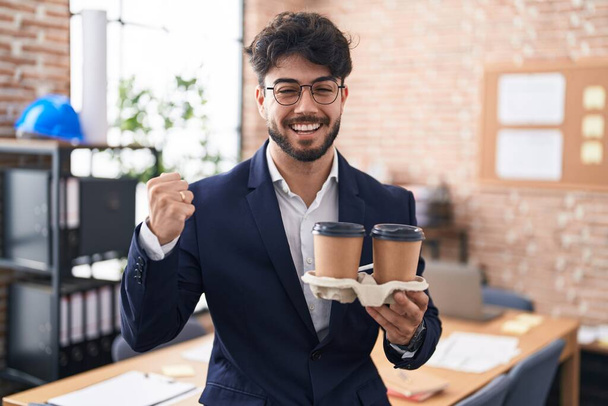 Ισπανόφωνος άντρας με μούσι που δουλεύει στο γραφείο κρατώντας τον καφέ ουρλιάζοντας περήφανος, γιορτάζοντας τη νίκη και την επιτυχία πολύ ενθουσιασμένος με το σηκωμένο χέρι  - Φωτογραφία, εικόνα