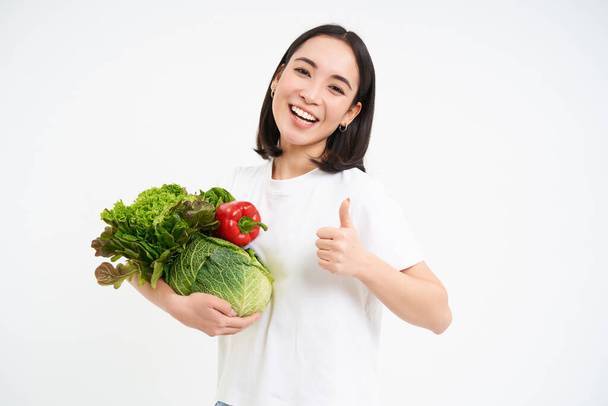 Lachende gelukkig aziatische vrouw toont duimen omhoog en groenten, het eten van gezond rauw voedsel, geïsoleerd over witte achtergrond. - Foto, afbeelding