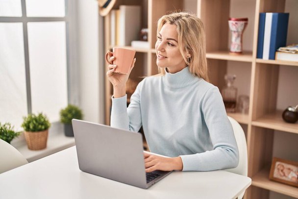 Молода блондинка використовує ноутбук і п'є каву, сидячи на столі вдома
 - Фото, зображення