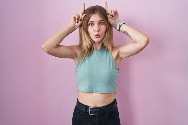 Blonde caucasian woman standing over pink background doing funny gesture with finger over head as bull horns  - Фото, зображення