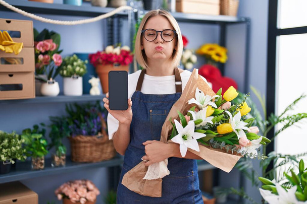 スマートフォンの画面を見せている花屋で働く若い白人女性は面白い顔で頬をむいています。空気を吸い込み空気を吸い込み.  - 写真・画像