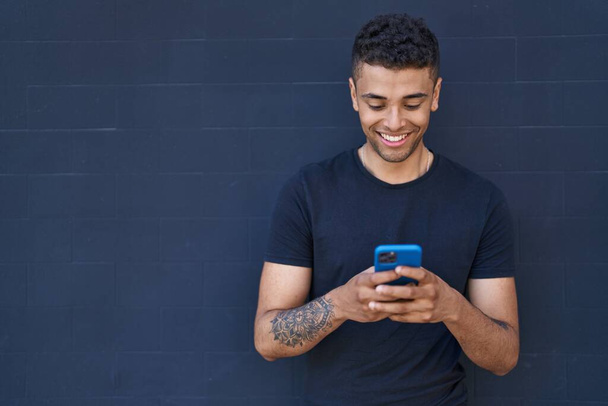 Αφροαμερικανός άντρας χαμογελά με αυτοπεποίθηση χρησιμοποιώντας smartphone πάνω από μαύρο φόντο - Φωτογραφία, εικόνα