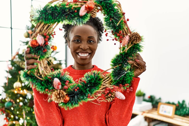 Αφροαμερικανή γυναίκα χαμογελά αυτοπεποίθηση κρατώντας χριστουγεννιάτικα στεφάνια διακόσμηση στο σπίτι - Φωτογραφία, εικόνα