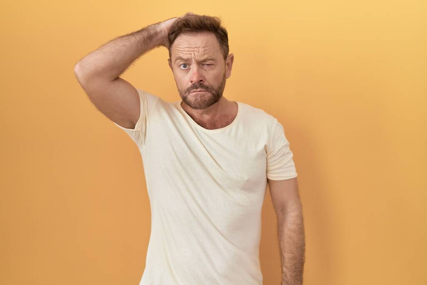 Middle age man with beard standing over yellow background confuse and wondering about question. uncertain with doubt, thinking with hand on head. pensive concept.  - Photo, Image