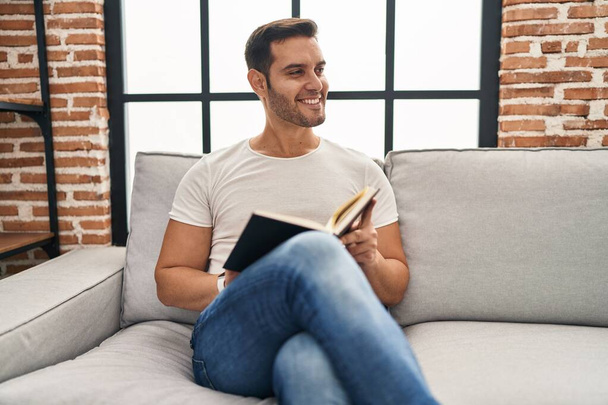 Young hispanic man reading book sitting on sofa at home - Photo, Image