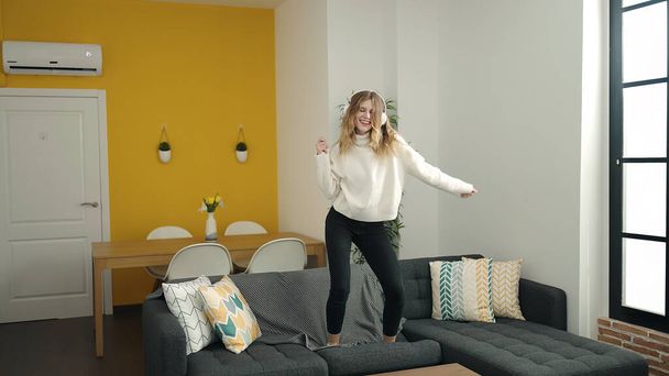 Nuori blondi nainen seisoo sohvalla kuunnellen musiikkia tanssia kotona - Valokuva, kuva