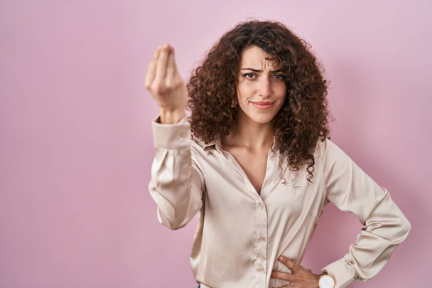 Hiszpanka z kręconymi włosami stojąca nad różowym tłem wykonująca włoski gest ręką i palcami  - Zdjęcie, obraz