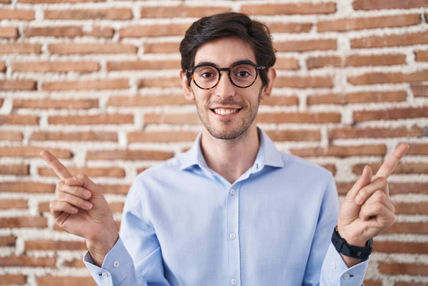 Fiatal spanyol férfi áll téglafal háttér mosolygós magabiztos mutató ujjaival különböző irányokba. hirdetési hely másolása  - Fotó, kép