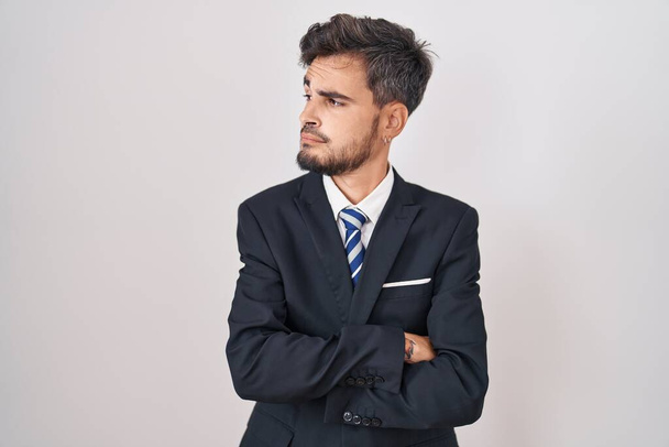 Jeune homme hispanique avec des tatouages portant un costume d'affaires et une cravate regardant sur le côté avec les bras croisés convaincu et confiant  - Photo, image