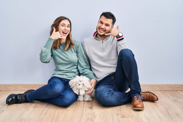 Молодая латиноамериканская пара, сидящая на полу с собакой, улыбающейся, делая телефонный жест рукой и пальцами, как будто разговаривая по телефону. коммуникационные концепции.  - Фото, изображение