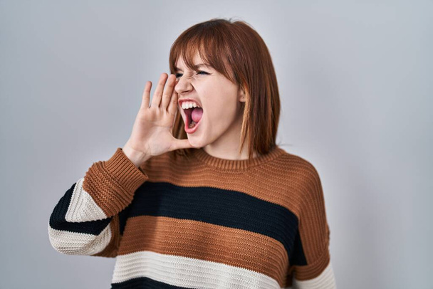 Young beautiful woman wearing striped sweater over isolated background shouting and screaming loud to side with hand on mouth. communication concept.  - Foto, immagini