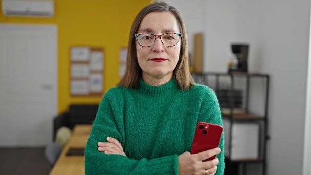 Mujer hispana madura con trabajador de negocios de cabello gris con cara seria sosteniendo teléfono inteligente en la oficina - Foto, Imagen