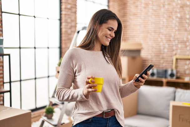Young beautiful hispanic woman using smartphone drinking coffee at new home - Zdjęcie, obraz