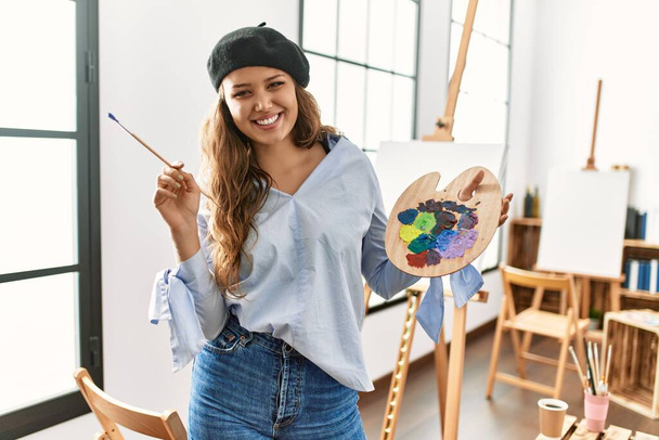 Mladá krásná hispánská žena umělec usmívá sebevědomě drží štětec a paletu v ateliéru umění - Fotografie, Obrázek