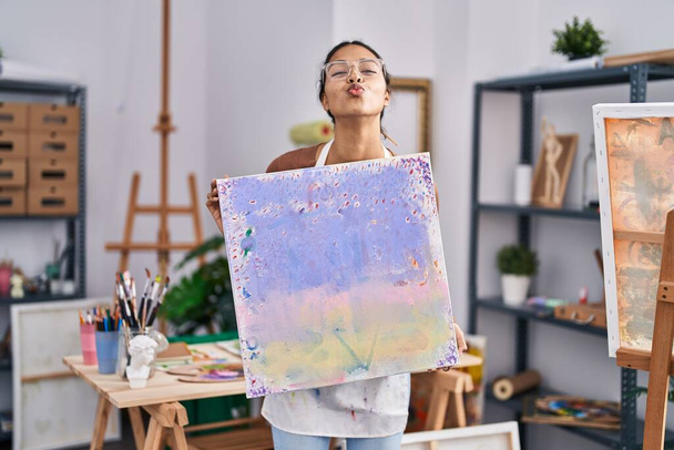 Young brazilian woman holding painter canvas at art studio looking at the camera blowing a kiss being lovely and sexy. love expression.  - Photo, Image
