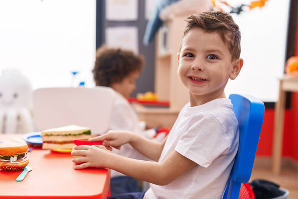 幼稚園のテーブルの上に座って食べ物おもちゃで遊んでいる大人の男の子 - 写真・画像
