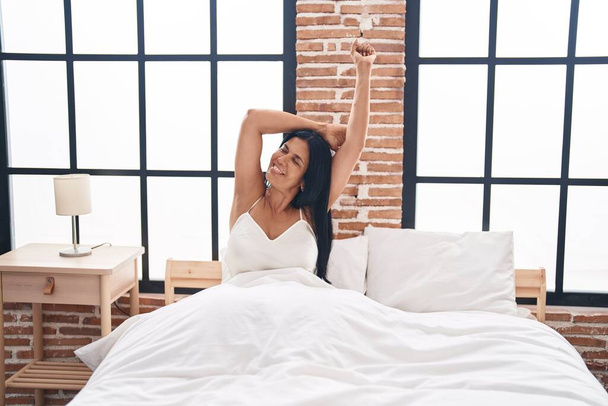 Middle age hispanic woman waking up stretching arms at bedroom - Foto, afbeelding