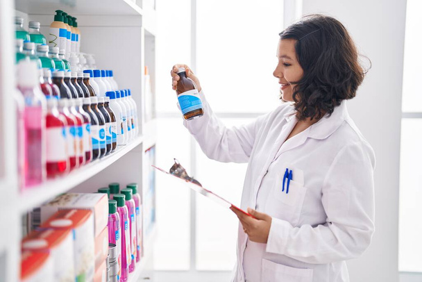 Young woman pharmacist holding medication bottle reading document at pharmacy - 写真・画像