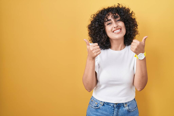 Young middle east woman standing over yellow background success sign doing positive gesture with hand, thumbs up smiling and happy. cheerful expression and winner gesture.  - Photo, Image