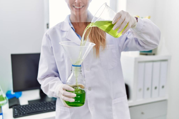 Laboratuvarda bilim adamı üniforması giyen genç sarışın kadın sıvıyı ölçüyor. - Fotoğraf, Görsel