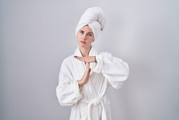 Blonde caucasian woman wearing bathrobe doing time out gesture with hands, frustrated and serious face  - 写真・画像