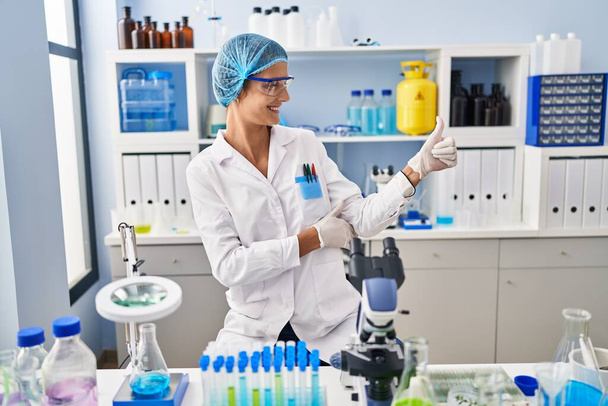 Brunette woman working at scientist laboratory looking proud, smiling doing thumbs up gesture to the side  - Foto, Bild
