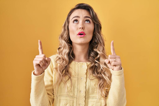 Young caucasian woman standing over yellow background amazed and surprised looking up and pointing with fingers and raised arms.  - Photo, Image