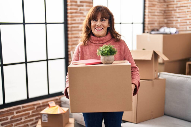 Middelbare leeftijd Spaanse vrouw verhuizen naar een nieuw huis met kartonnen doos glimlachend met een vrolijke en koele glimlach op het gezicht. tanden tonen.  - Foto, afbeelding