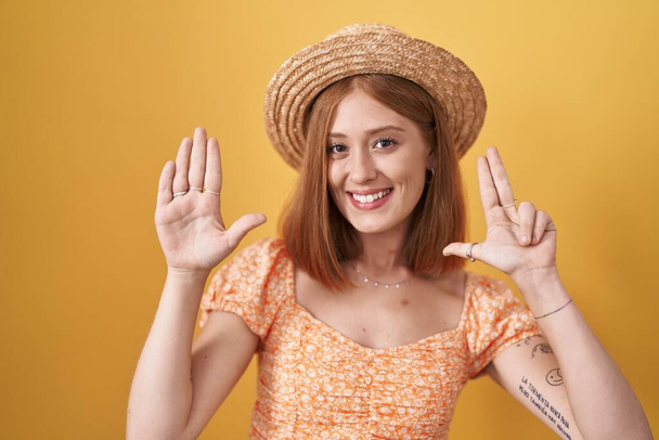 Young redhead woman standing over yellow background wearing summer hat showing and pointing up with fingers number eight while smiling confident and happy.  - Photo, Image